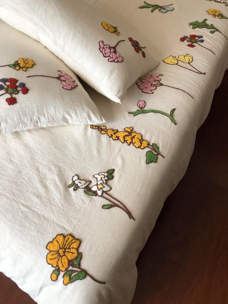 K&ouml;nnte beinhalten: Cremefarbenes Bettw&auml;sche-Set mit gestickten Blumenmustern. Das Set beinhaltet einen Bettbezug und zwei Kissenbez&uuml;ge, verziert mit bunten gestickten Blumen in Gelb, Rosa, Rot und Blau. Die Blumen sind &uuml;ber den Stoff verteilt.