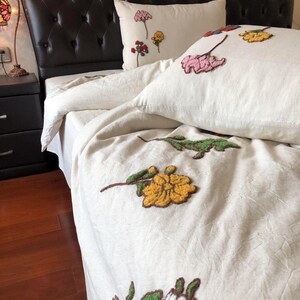 K&ouml;nnte beinhalten: Cremefarbenes Bettw&auml;sche-Set mit gestickten Blumendesigns in Rosa, Gelb und Gr&uuml;n. Das Set beinhaltet einen Bettbezug und Kissenbez&uuml;ge. Das Bett hat ein dunkelbraunes, getuftetes Kopfteil.