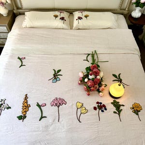 K&ouml;nnte beinhalten: Ein cremefarbenes Bettw&auml;sche-Set mit gestickten Blumenmustern. Das Set umfasst zwei Kissenbez&uuml;ge und eine Bettdecke, die mit verschiedenen bunten Blumenmotiven verziert sind. Ein Strau&szlig; aus rosa und roten Rosen liegt auf dem Bett, zusammen mit einer kleinen gelben Lampe.