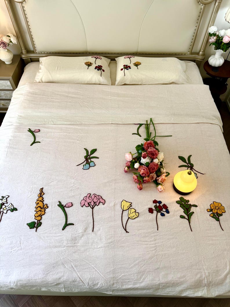 K&ouml;nnte beinhalten: Ein cremefarbenes Bettw&auml;sche-Set mit gestickten Blumenmustern. Das Set umfasst zwei Kissenbez&uuml;ge und eine Bettdecke, die mit verschiedenen bunten Blumenmotiven verziert sind. Ein Strau&szlig; aus rosa und roten Rosen liegt auf dem Bett, zusammen mit einer kleinen gelben Lampe.