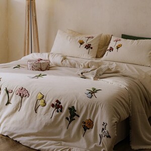 K&ouml;nnte beinhalten: Cremefarbenes Bettw&auml;sche-Set mit gestickten Blumenmustern in verschiedenen Farben. Das Set beinhaltet einen Bettbezug und Kissenbez&uuml;ge. Eine dekorative Geschenkbox mit floralem Muster liegt auf dem Bett, zusammen mit einer gefalteten Decke.