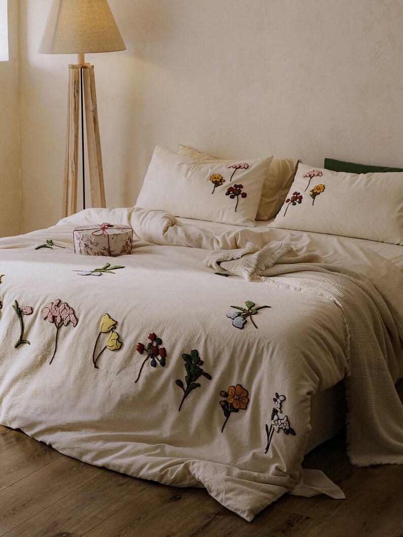 K&ouml;nnte beinhalten: Cremefarbenes Bettw&auml;sche-Set mit gestickten Blumenmustern in verschiedenen Farben. Das Set beinhaltet einen Bettbezug und Kissenbez&uuml;ge. Eine dekorative Geschenkbox mit floralem Muster liegt auf dem Bett, zusammen mit einer gefalteten Decke.