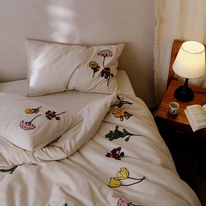 K&ouml;nnte beinhalten: Cremefarbenes Bettw&auml;sche-Set mit gestickten Blumenmustern in verschiedenen Farben. Das Set umfasst ein Kissen und einen Bettbezug mit rosa, gelben, blauen und roten Blumen. Eine kleine Lampe und ein Buch sind auf einem Nachttisch aus Holz zu sehen.