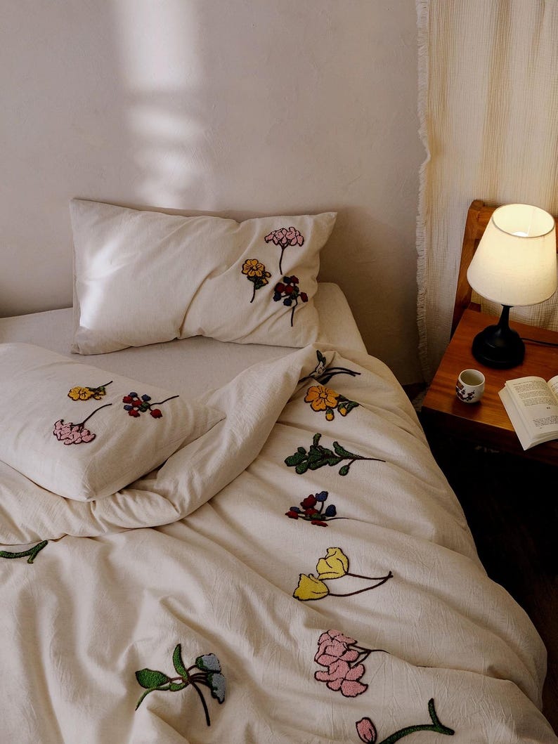 K&ouml;nnte beinhalten: Cremefarbenes Bettw&auml;sche-Set mit gestickten Blumenmustern in verschiedenen Farben. Das Set umfasst ein Kissen und einen Bettbezug mit rosa, gelben, blauen und roten Blumen. Eine kleine Lampe und ein Buch sind auf einem Nachttisch aus Holz zu sehen.