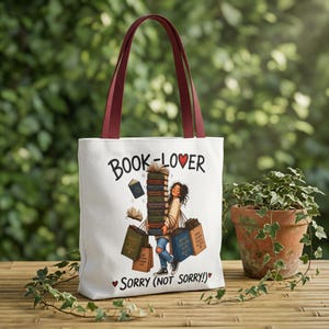May include: White tote bag with claret-coloured handles. The bag features the text "BOOK-LOVER" and "SORRY (NOT SORRY!)", along with an illustration of a person carrying books and shopping bags. The background shows natural greenery.