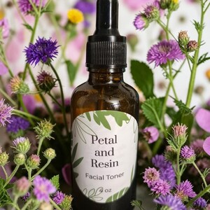 May include: A brown glass bottle with a black dropper cap containing facial toner. The white label features green leaf illustrations and the text Petal and Resin Facial Toner 4 oz. The bottle is surrounded by purple and yellow wildflowers.