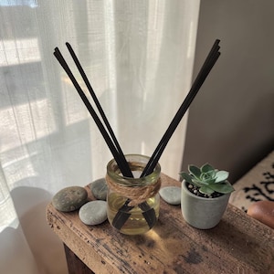 May include: A glass jar diffuser with black reeds, filled with a yellow liquid, and wrapped with twine. The jar sits on a wooden surface with smooth stones and a small succulent in a gray pot. The background includes a white curtain.