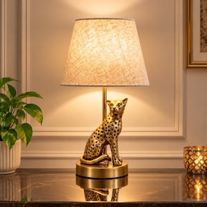 May include: A table lamp featuring a gold-coloured leopard sculpture as the base, topped with a tapered, textured off-white fabric lampshade. The lamp emits a warm glow, illuminating the metallic finish of the leopard and the circular base.