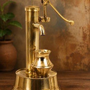 Könnte beinhalten: Eine Wasserpumpe aus Messing mit einer kleinen passenden Vase. Die Pumpe hat einen Griff und einen Ausguss, und die Vase steht auf einem runden Sockel. Pumpe und Vase sind goldfarben und haben eine polierte Oberfläche. Der Hintergrund ist verschwommen braun.