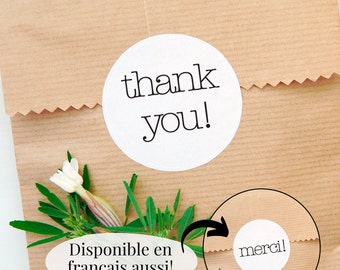 Op de afbeelding: Een bruine papieren zak met een witte ronde sticker waarop "thank you!" staat. Er is ook een kleinere witte ronde sticker op de zak met de tekst "merci!" De tekst "Disponible en français aussi!" staat op een witte ovale sticker op de zak.