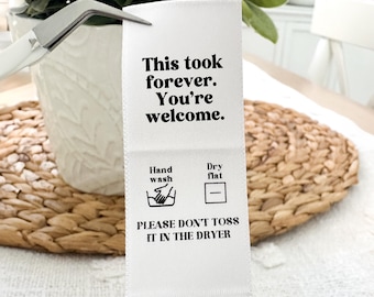 May include: White fabric care label with black text and icons. The text reads "This took forever. You're welcome." and "PLEASE DON'T TOSS IT IN THE DRYER". The icons indicate hand wash and dry flat.