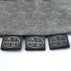 May include: Three black fabric labels with the word "labelicious" in a script font. Each label has a white oval border with laundry symbols. The labels are attached to a grey fabric.