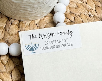 May include: A white rectangular address label featuring the text "The Wilson Family" in a handwritten font. Below the name is a menorah graphic and address details. The label is on a woven surface.