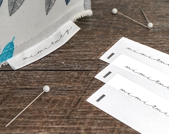 May include: A close-up of a white fabric with a blue and gray feather pattern. The fabric has a label that reads "mimi+meg". There are three white cards with the text "mimi+meg" written on them. The cards are on a wooden surface with white pins.