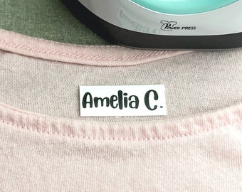 May include: A white iron-on label with black text that reads "Amelia C." The label is on a pink shirt.