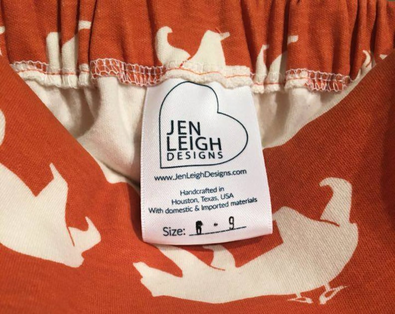 Pode incluir: Tecido laranja com um padr&atilde;o branco de p&aacute;ssaros. Uma etiqueta branca com o texto "Jen Leigh Designs" e o endere&ccedil;o do site "www.JenLeighDesigns.com". A etiqueta tamb&eacute;m diz "Handcrafted in Houston, Texas, USA" e "With domestic & imported materials". A etiqueta inclui o tamanho "6 - 9".