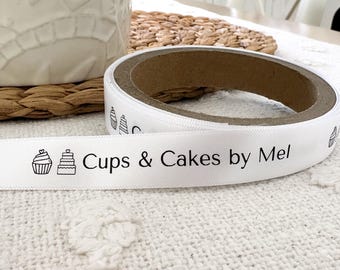 May include: White satin ribbon with black ink printing. The ribbon has a cupcake and a layered cake design on either end. The text on the ribbon reads "Cups & Cakes by Mel".