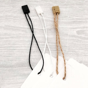 Puede incluir: Tres tipos diferentes de etiquetas de cuerda con puntas de plástico. Las etiquetas son negras, blancas y marrones. Las etiquetas están unidas a papel blanco.