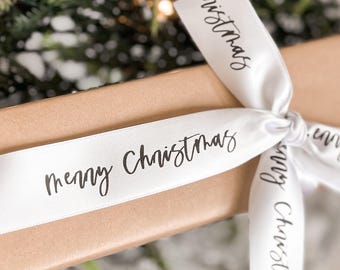 May include: White satin ribbon with the text "Merry Christmas" printed in black. The ribbon is tied around a brown gift box.