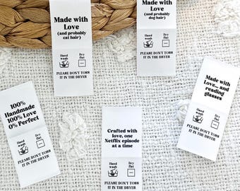 May include: Five white fabric labels with black text and icons. The labels say "Made with Love (and probably cat hair)", "Made with Love (and probably dog hair)", "Made with Love... and reading glasses", "Crafted with love, one Netflix episode at a time", and "100% Handmade 100% Love 0% Perfect". Each label has icons for hand wash and dry flat, and the text "Please don't toss it in the dryer".
