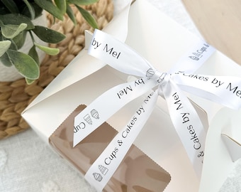 May include: White gift box with a clear window, tied with a white ribbon that says "Cups & Cakes by Mel" in black text.