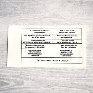 以下が含まれることがあります： 「Ne pas enlever avant la livraison au consommateur」と「Not to be removed until delivered to the consumer」という黒いテキストが書かれた白いラベル。「Cet article contient des MATERIAUX NEUFS SEULEMENT」と「This article contains NEW MATERIAL ONLY」も記載されている。また、「Fabriquée par Glup bébé Inc No. de permis: QC-009588」と「Made by Glup bébé Inc Reg. No.: QC-009588」も記載されている。さらに、「CONTENU INTERIEUR: 100% POLYESTER CONTENU EXTERIEUR: 100% COTON」と「INTERIOR CONTENT: 100% POLYESTER EXTERIOR CONTENT: 100% COTTON」も記載されている。ラベルには「FAIT AU CANADA / MADE IN CANADA」とも書かれている。