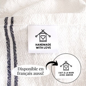 May include: White fabric with a black and white label that says "Handmade with Love". The label has a small icon of a heart and knitting needles. The fabric has two black stripes. The text "Disponible en français aussi!" is written in a white oval on the fabric. A white circle with a black and white label that says "Fait à la main avec amour" is shown next to the oval.