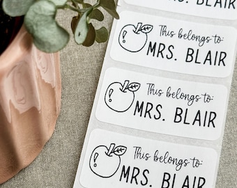 May include: Four white rectangular labels with a black and white illustration of an apple with a leaf and an exclamation mark. Each label has the text "This belongs to: MRS. BLAIR" in black.
