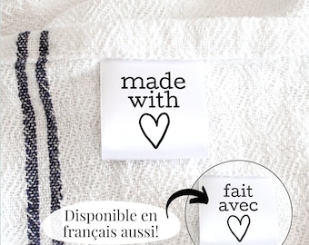 May include: White fabric label with black text that reads "made with" and a heart. The label is attached to a white fabric with blue stripes. Below the label is a white oval with black text that reads "Disponible en français aussi!" and an arrow pointing to a second white fabric label with black text that reads "fait avec" and a heart.
