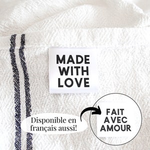May include: White fabric with a black and white label that reads "MADE WITH LOVE". The label is attached to the fabric with a white thread. Below the label is a white oval with black text that reads "Disponible en français aussi!" and a black arrow pointing to a white circle with black text that reads "FAIT AVEC AMOUR".