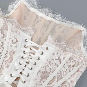 May include: Close-up of a white lace corset with a delicate floral pattern. The corset features a lace-up front with silver eyelets and a scalloped lace trim along the neckline. The fabric underneath is a nude color.