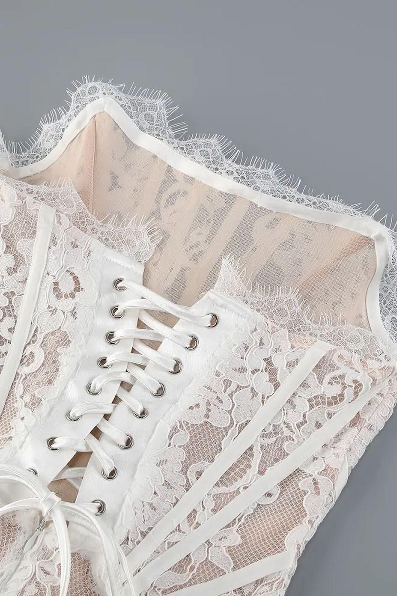 May include: Close-up of a white lace corset with a delicate floral pattern. The corset features a lace-up front with silver eyelets and a scalloped lace trim along the neckline. The fabric underneath is a nude color.