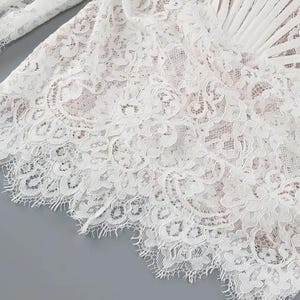 May include: Close-up of white lace fabric with intricate floral patterns. The delicate lace is layered, creating depth and texture. The edges are scalloped, adding a refined finish. The fabric appears to be part of a garment.