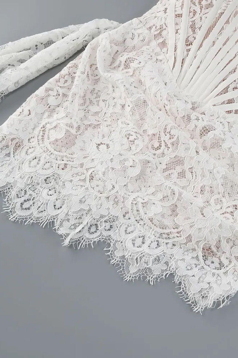 May include: Close-up of white lace fabric with intricate floral patterns. The delicate lace is layered, creating depth and texture. The edges are scalloped, adding a refined finish. The fabric appears to be part of a garment.