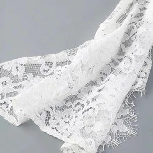 May include: Close-up of white lace fabric with a floral pattern. The delicate material is draped, revealing intricate details and textures. The lace has a scalloped edge and is set against a gray background.