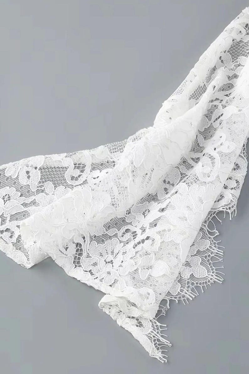 May include: Close-up of white lace fabric with a floral pattern. The delicate material is draped, revealing intricate details and textures. The lace has a scalloped edge and is set against a gray background.