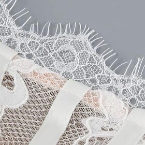 May include: Close-up of white lace fabric with a delicate floral pattern and scalloped edges. The image shows a section of a garment, possibly lingerie or a dress, with satin straps. The background is a solid gray.