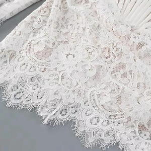 May include: Close-up of a white lace fabric with intricate floral patterns. The delicate lace has a scalloped edge and is layered over a neutral-toned fabric. The image highlights the texture and detail of the lace, suggesting a garment or textile.