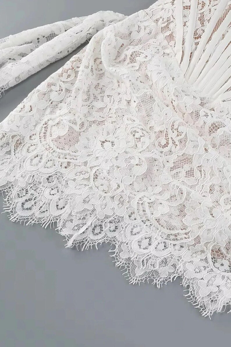 May include: Close-up of a white lace fabric with intricate floral patterns. The delicate lace has a scalloped edge and is layered over a neutral-toned fabric. The image highlights the texture and detail of the lace, suggesting a garment or textile.