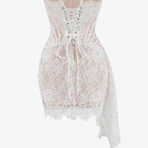 May include: A white lace corset dress with a sweetheart neckline and a lace-up back. The dress features a fitted bodice and a short skirt with a flowing, asymmetrical lace overlay. The dress is a delicate and romantic piece.
