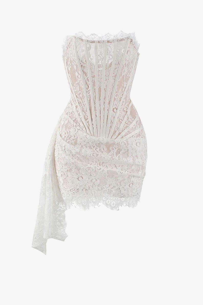 May include: A white lace dress with a strapless, corset-style bodice. The dress features a draped skirt and a delicate lace trim along the neckline and hem. The dress is a formal wear item.