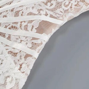 May include: Close-up of a white lace garment with a delicate floral pattern. The garment features horizontal white satin ribbons that add structure and detail to the design. The background is a solid gray.