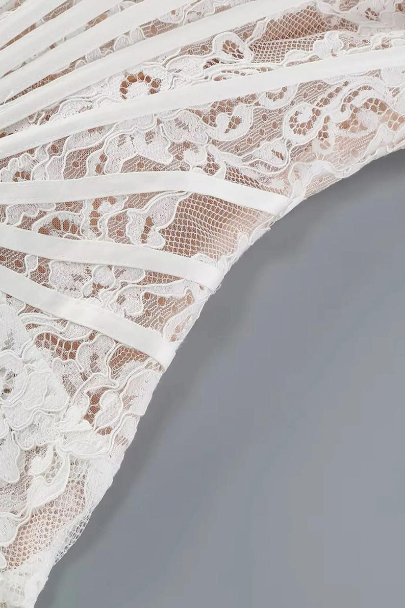 May include: Close-up of a white lace garment with a delicate floral pattern. The garment features horizontal white satin ribbons that add structure and detail to the design. The background is a solid gray.