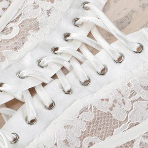 May include: Close-up of a white lace corset with satin ribbon lacing. The corset features intricate floral lace detailing and silver eyelets. The background is a light beige color.
