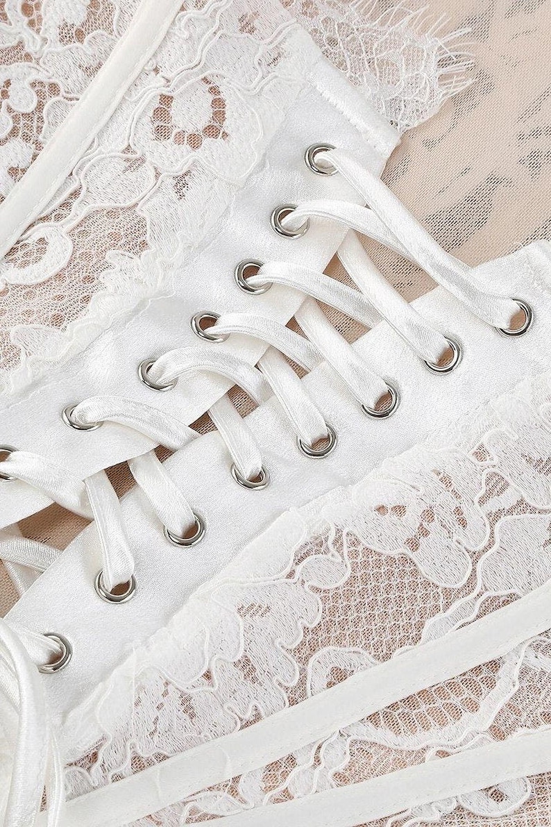 May include: Close-up of a white lace corset with satin ribbon lacing. The corset features intricate floral lace detailing and silver eyelets. The background is a light beige color.