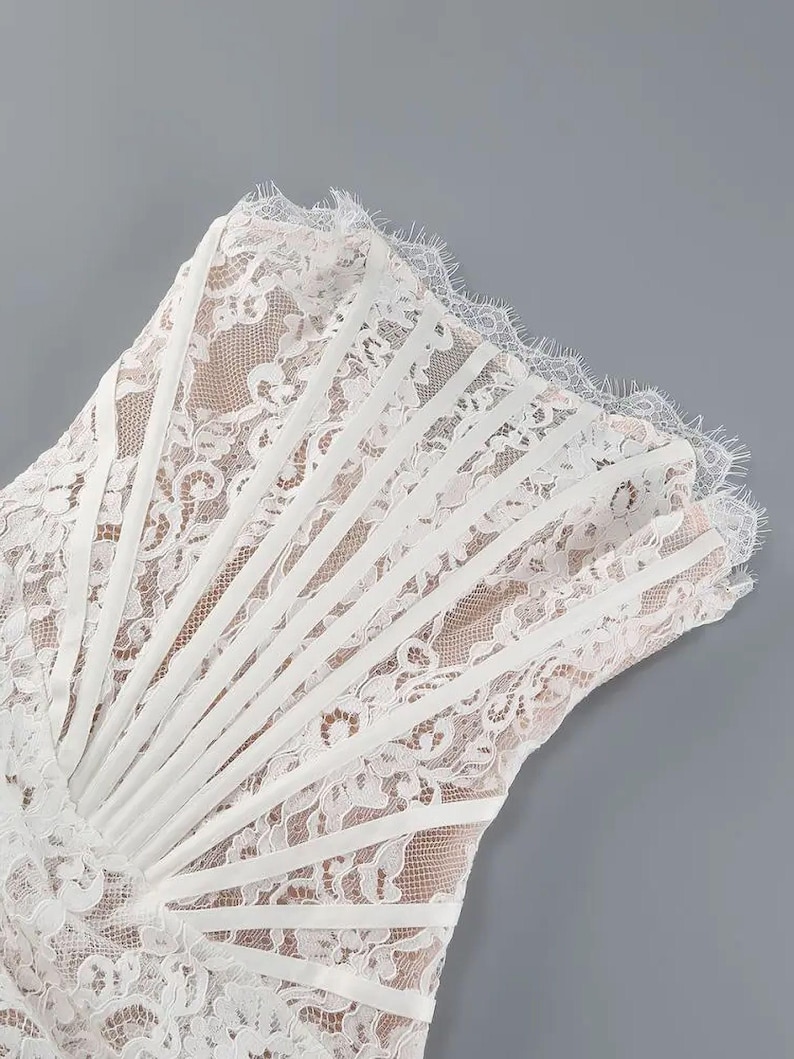 May include: White lace dress with a detailed floral pattern. The dress features a strapless design with vertical satin ribbon accents and a scalloped lace trim. The dress is set against a gray background.
