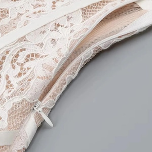 May include: Close-up of a white lace dress with a zipper detail. The dress features intricate floral lace patterns and a beige lining visible through the delicate fabric. The zipper is silver.