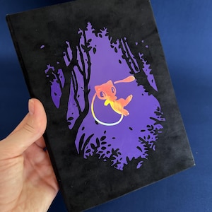May include: A black rectangular notebook featuring a cutout design on the front cover. The cutout reveals a purple background with a silhouette of trees and a small, light-colored creature with a long tail floating in the center of the scene.