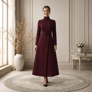 May include: A long-sleeved, floor-length coat in a deep burgundy color. The coat features a high mock neck collar, a fitted bodice, and a matching fabric belt with a rectangular buckle at the waist. The skirt has a clean, straight-cut silhouette.