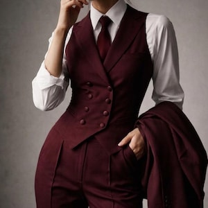 May include: A deep burgundy double-breasted waistcoat with matching tailored trousers and a white long-sleeved button-down shirt. A matching burgundy necktie is worn with the shirt, and a coordinating burgundy blazer is held in one hand.